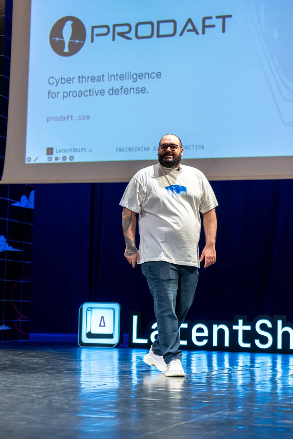 Alican Kiraz smiles as he walks across the LatentShift stage in a white printed T-shirt beneath a projected PRODAFT slide.