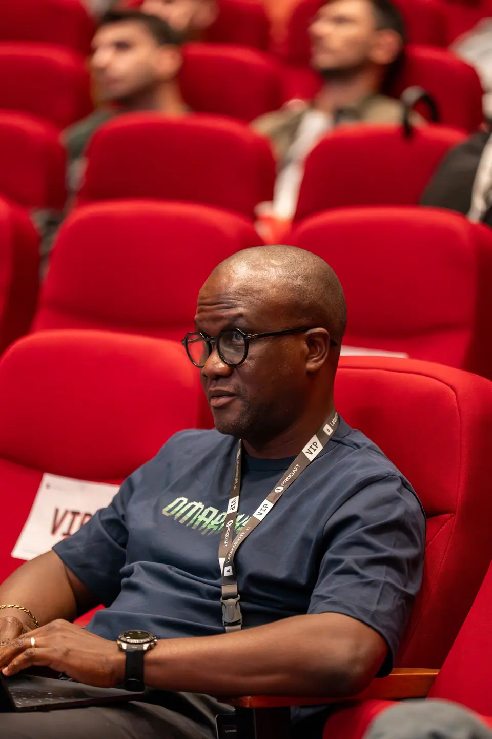 An attendee wearing round glasses and a VIP lanyard sits in a red auditorium seat with a laptop on their lap.