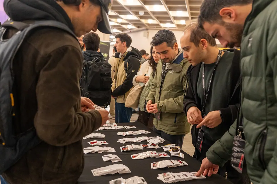Attendees lean over a black-covered table to examine rows of small packaged items, while one visitor holds a packet.