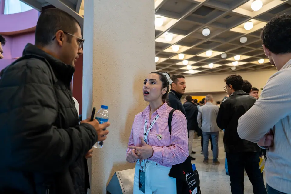 Bilge Gündüz in a pink striped shirt talks with a man holding a water bottle beside a pillar in the busy foyer.