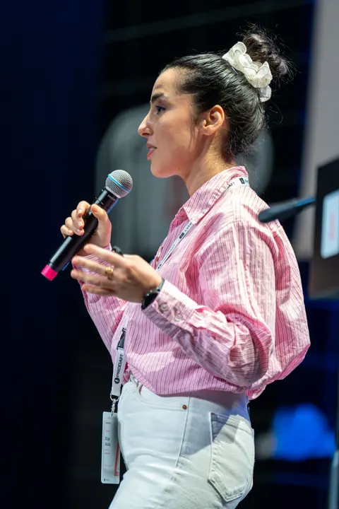 Bilge Gündüz in a pink striped shirt speaks into a handheld microphone and gestures with her free hand on stage.