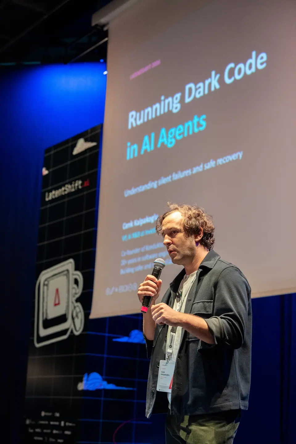 Cenk Kalpakoglu in a dark jacket holds a microphone beneath a slide titled “Running Dark Code in AI Agents.”