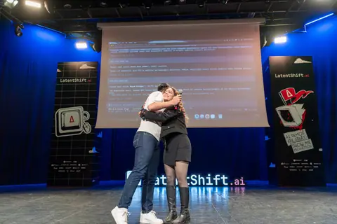 Two people embrace at center stage beneath a projected screen, framed by blue curtains and LatentShift banners.