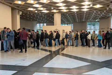 Attendees queue across a spacious foyer with a patterned stone floor and rows of round ceiling lights.