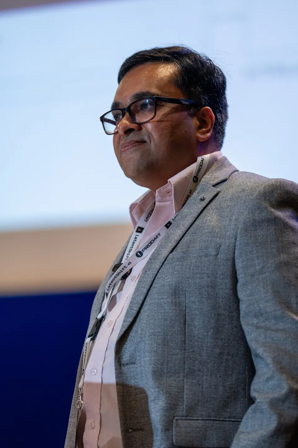 Close-up of Jigar Mody wearing black glasses, a gray blazer and a pale pink shirt in front of the projection screen.