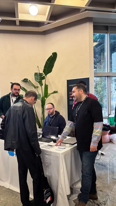A man points to brochures on a booth table as a visitor in a leather jacket leans forward beside open laptops.