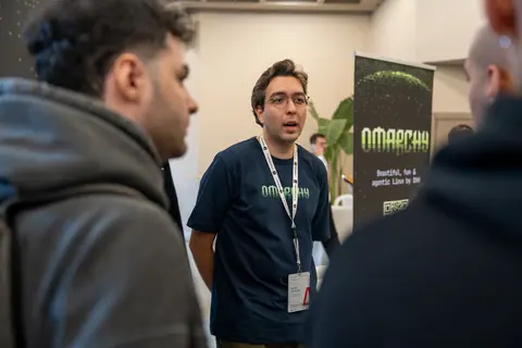 An attendee wearing round glasses and a navy T-shirt speaks with two people beside a tall display banner.
