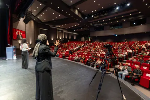 A woman in a patterned headscarf gestures onstage beside another woman facing the seated audience, with a tripod-mounted camera nearby.