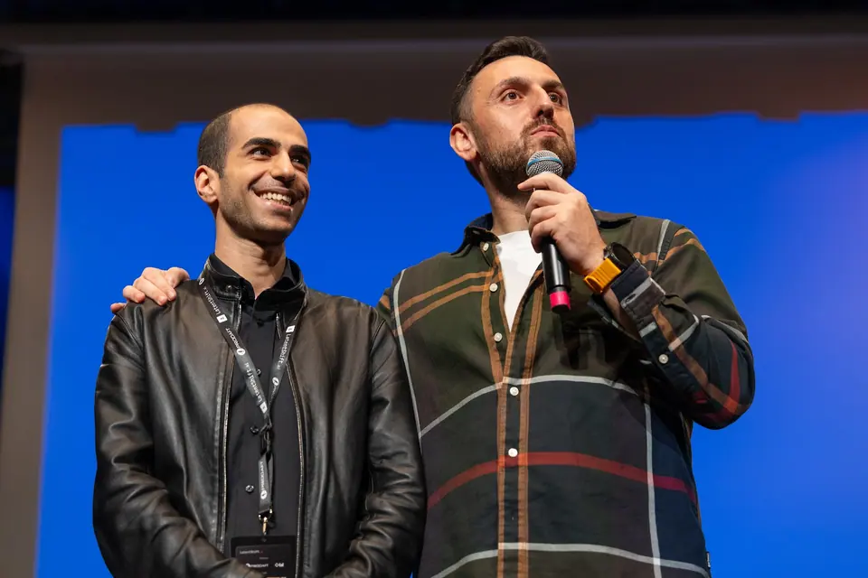 A man in a plaid shirt holds a microphone and rests his arm around a smiling man in a black leather jacket.