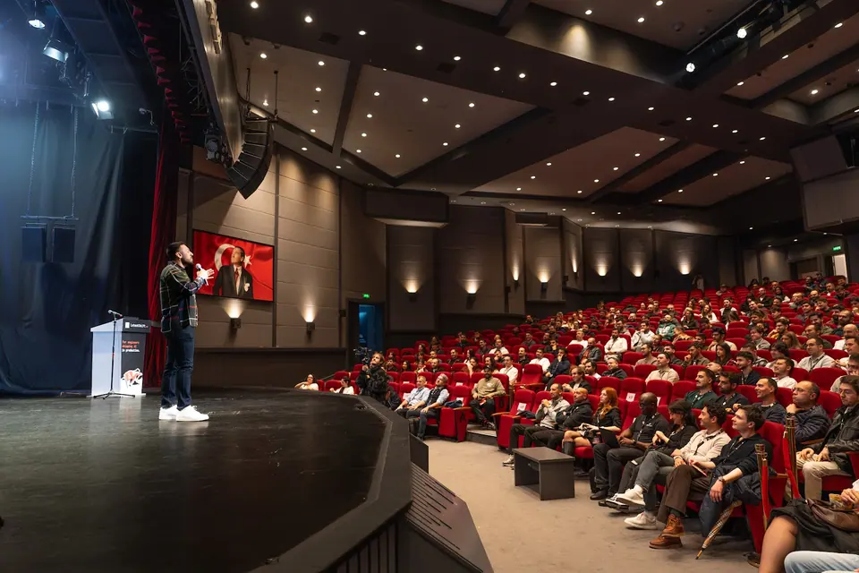 A speaker in a dark plaid shirt holds a microphone and gestures toward the audience seated in red auditorium chairs.
