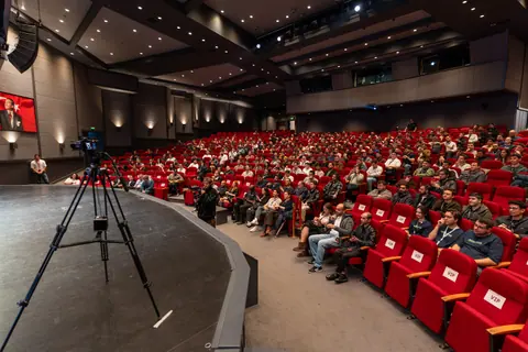 Attendees fill rows of red auditorium seats facing the stage, with a camera on a tripod in the foreground.