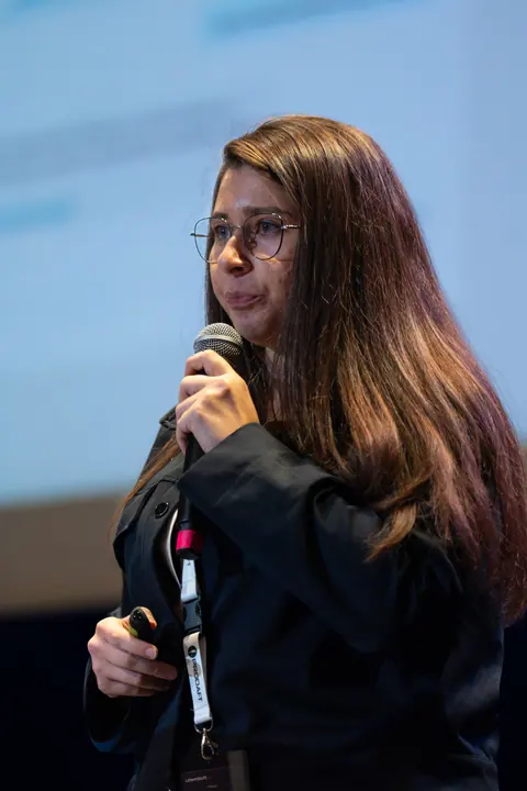 Close view of Şeyma Sarıgil wearing round glasses and a black jacket, holding a microphone and presentation clicker.