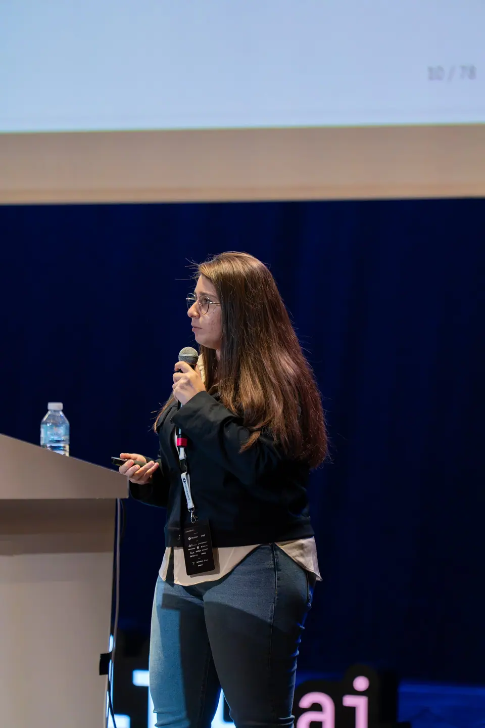 Seen in profile beside the lectern, Şeyma Sarıgil with long hair and glasses holds a microphone and a presentation remote.