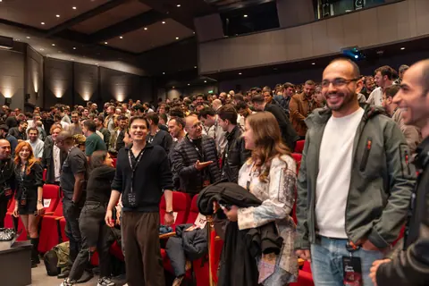 Smiling attendees stand and talk between rows of red auditorium seats, with several facing the camera.