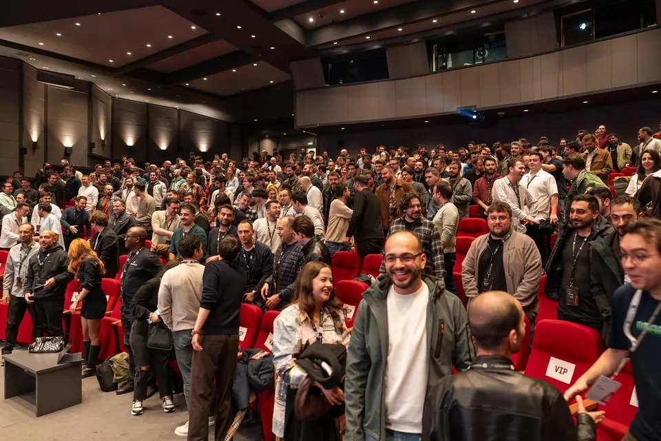 A smiling attendee in a green jacket stands beside an attendee holding a black coat, as people gather between rows of red auditorium seats.