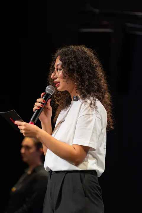 A curly-haired speaker wearing glasses and a white T-shirt reads from a card while holding a microphone on stage.