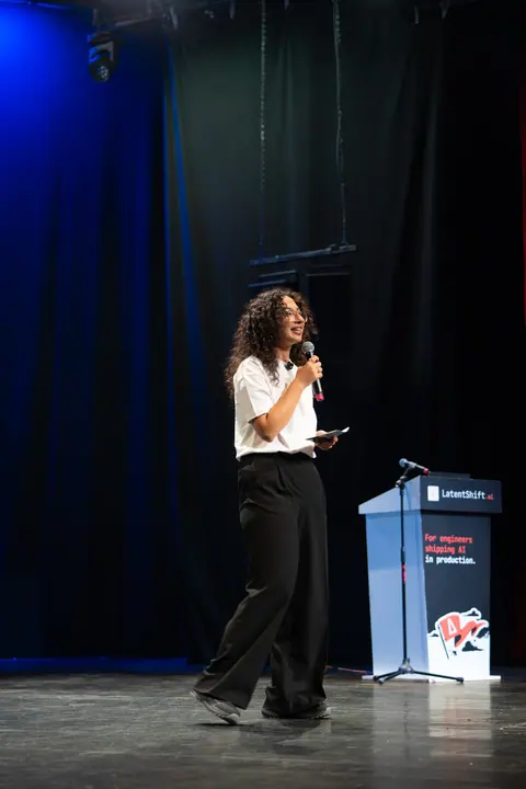 A speaker in a white T-shirt and black trousers holds a microphone and notes beside the LatentShift lectern on stage.