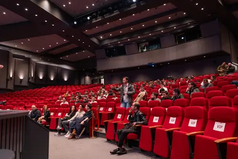 An attendee in a dark jacket stands among red auditorium seats and gestures toward the stage while nearby attendees remain seated.