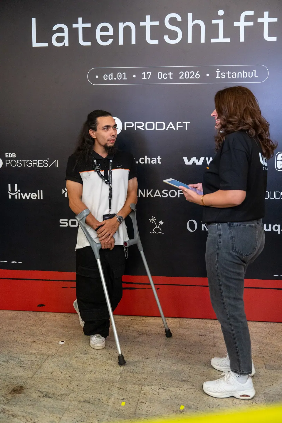 Two attendees face each other at the LatentShift backdrop; one holds a tablet while the other stands with forearm crutches.