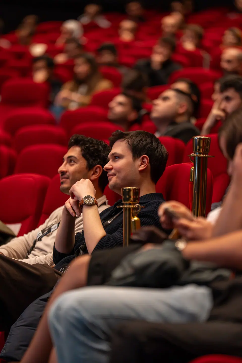 Two attendees smile from adjacent red seats, with gold-colored barrier posts and more seated audience members around them.