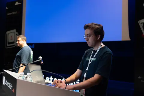A man with glasses types on a laptop at the LatentShift lectern while another man stands behind him beneath a blue screen.