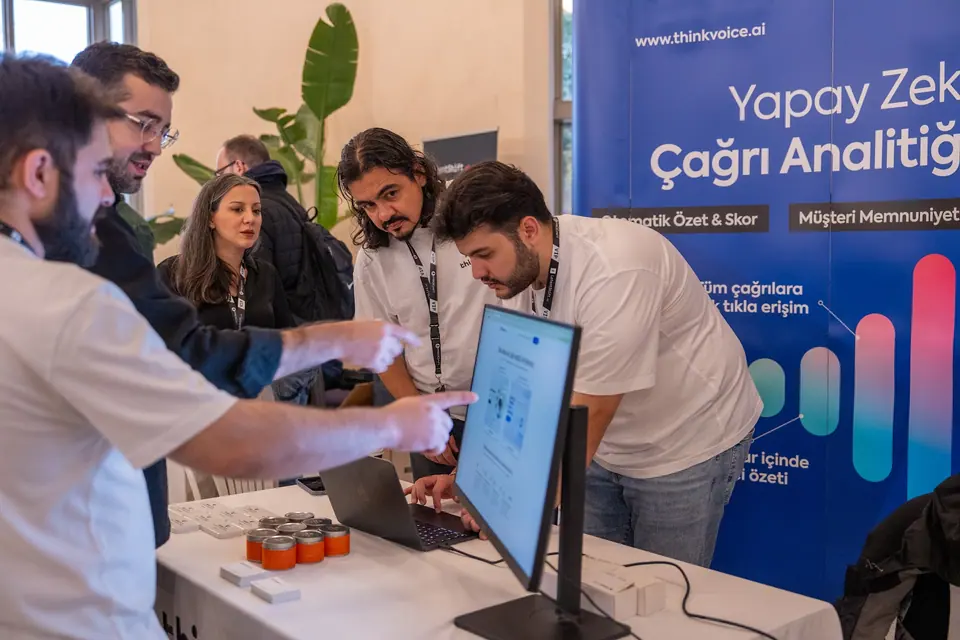 Visitors point toward a monitor as a man operates a laptop at a table in front of the ThinkVoice banner.