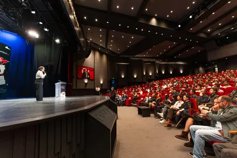 A wide view of the auditorium shows a speaker in a white blouse onstage facing rows of attendees in red seats.