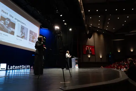 A woman stands beside the lectern while another gestures near a projected organizers slide, with seated attendees visible at right.