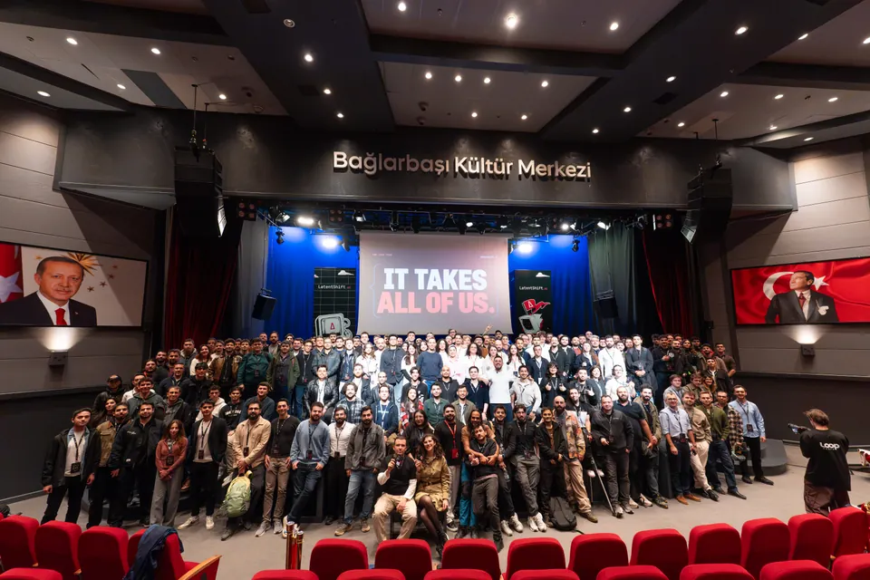 A large group poses in front of the LatentShift stage beneath a screen reading “IT TAKES ALL OF US.”