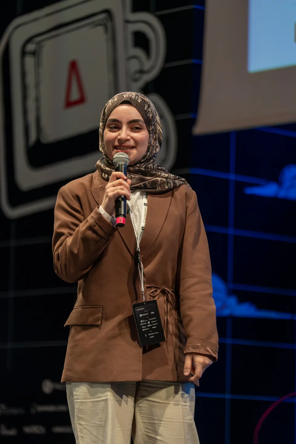 Ebrar Kızıloğlu smiles while holding a microphone near her mouth, with the LatentShift emblem behind her.