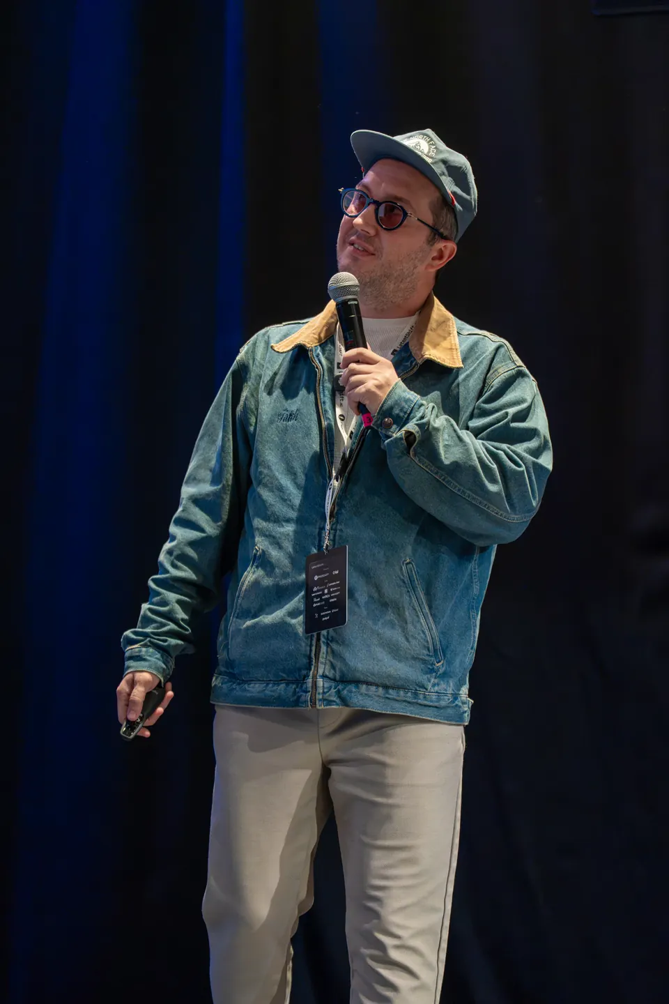 Fatih Kadir Akın in a denim jacket and cap looks upward while holding a microphone near their mouth and a presentation remote at their side.