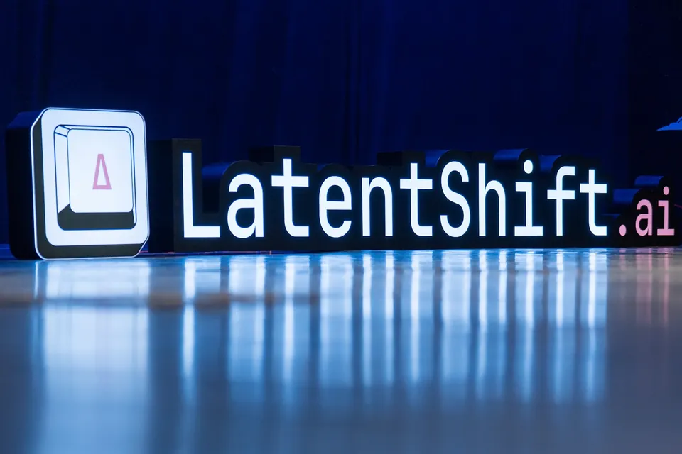 An illuminated LatentShift.ai sign and logo reflect across the polished stage floor in front of a blue curtain.