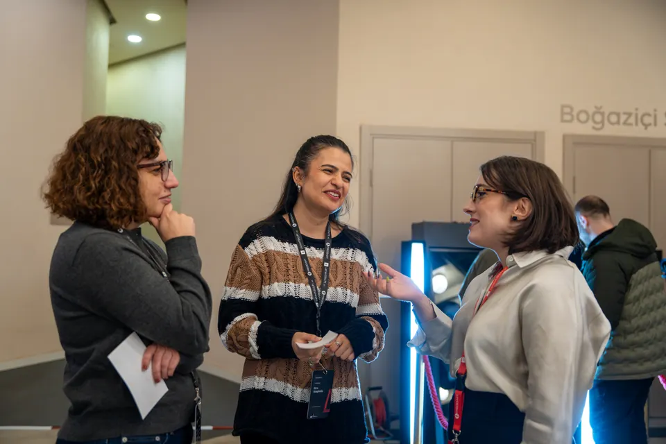 Three women chat in the foyer, with one gesturing, another smiling in a striped sweater, and the third listening with her hand at her chin.